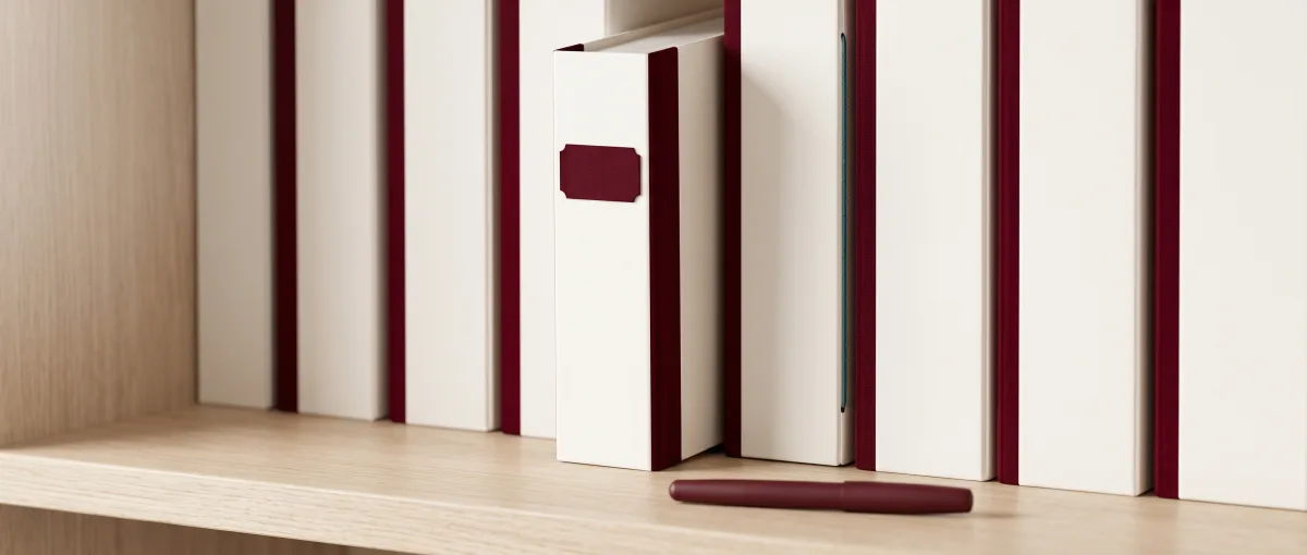 Rangée de classeurs crème à dos oxblood sur une étagère en chêne, l'un tiré en avant, un stylo posé devant